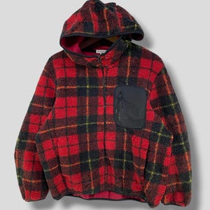 Può includere: Una giacca in pile con cappuccio a quadri rossi e neri con una tasca nera sul petto. La giacca ha una cerniera completa e una fodera interna rossa. Anche il cappuccio è foderato in rosso. La giacca è realizzata in materiale morbido e soffice.