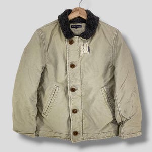 May include: Tan jacket with a dark gray faux fur collar and brown buttons. The jacket has two front pockets and a small patch near the top button. The brand name "BLUE BLUE" is visible on the collar.