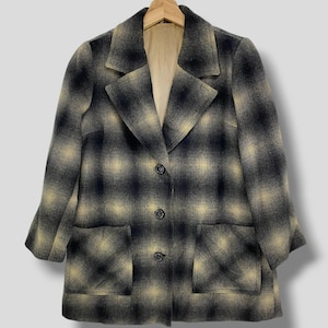 Può includere: Un cappotto vintage a quadri neri e beige. Il cappotto ha un design classico con colletto a revers, tre bottoni e due tasche anteriori. È appeso a una gruccia di legno. Il motivo del cappotto è a quadri grandi.