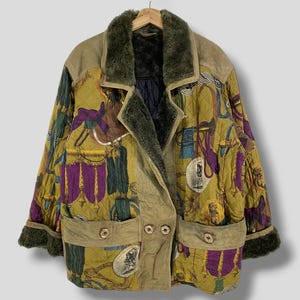 Pode incluir: Casaco acolchoado vintage com um padrão equestre único em tons de ouro, roxo e verde. O casaco apresenta gola e punhos de pele sintética, painéis semelhantes a camurça e bolsos abotoados. O casaco está pendurado num cabide de madeira.