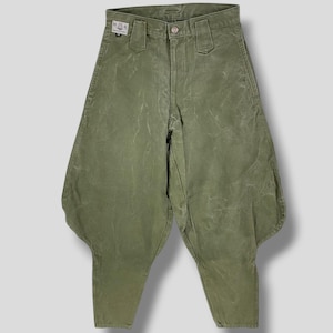 Könnte beinhalten: Olivgrüne Hose mit einem einzigartigen, ausgestellten Design. Die Hose hat einen Knopfverschluss und Gürtelschlaufen. Der Stoff hat ein leichtes Used-Aussehen. Ein kleines Etikett ist am Bund sichtbar. Die Hose wird vor einem neutralen Hintergrund gezeigt.