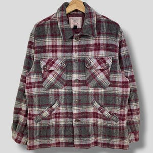 Puede incluir: Una camisa a cuadros en tonos grises, burdeos y crema. La camisa tiene dos bolsillos en el pecho y dos bolsillos inferiores. La camisa está colgada de una percha de madera. La etiqueta de la camisa dice "PROG".