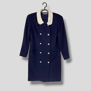 Puede incluir: Un vestido azul marino con cuello de encaje blanco y botones plateados. El vestido tiene cierre de doble botonadura y dos bolsillos.