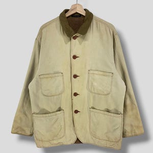 May include: A tan work jacket with a brown corduroy collar and four large front pockets. The jacket has brown buttons and is hanging on a wooden hanger. The jacket appears to be made of a durable, possibly canvas, material.