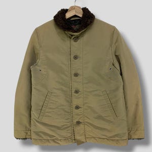 May include: A tan jacket with a brown faux fur collar, button closure, and two front pockets. The jacket is hanging on a wooden hanger against a plain background. The jacket appears to be made of a durable, possibly water-resistant material.