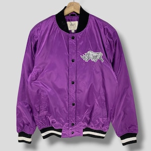 May include: A purple bomber jacket with a black collar and cuffs. The jacket features a rhino patch on the front and black buttons. The cuffs have a black and white striped design. The jacket is hanging on a wooden hanger.
