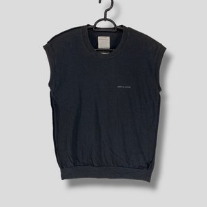 May include: Black sleeveless shirt with the text "COMME des GARÇONS" in small white letters. The shirt has a crew neck and a gathered waist. The garment is hanging on a black hanger.
