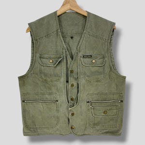 Peut inclure: Gilet vert olive avec plusieurs poches, dont deux poches zippées et deux poches à rabat boutonnées. Le gilet a une fermeture à boutons sur le devant et une petite étiquette qui indique "BRICK DAR". Le gilet est suspendu à un cintre en bois.