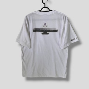 May include: White t-shirt featuring a black and gray graphic of an Xbox Kinect sensor on the back, with the Xbox logo above. The Xbox logo is also printed on the right sleeve.