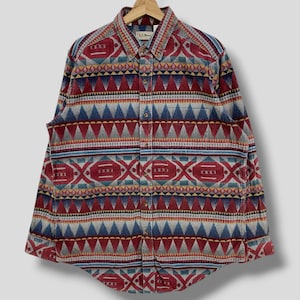 Puede incluir: Camisa abotonada con un colorido estampado geométrico. La camisa presenta una mezcla de triángulos, diamantes y otras formas rojas, azules, grises y amarillas. La camisa está colgada de una percha de madera.