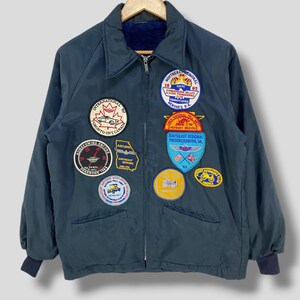 Könnte beinhalten: Dunkelgrüne Vintage-Jacke mit mehreren gestickten Aufnähern. Aufnäher mit Texten wie "Northeast Regional", "Toronto Ont Canada" und "Hershey Region". Die Jacke hat einen Reißverschluss, einen Kragen und marineblaue Bündchen.