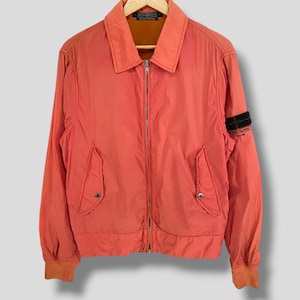 May include: A coral-colored zip-up jacket featuring a classic collar, long sleeves with ribbed cuffs, and two front pockets with snap closures. A black Stone Island patch is attached to the left sleeve, and the interior lining is a contrasting brown.