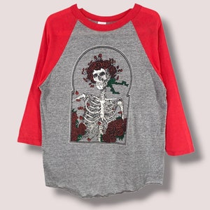 Puede incluir: Una camiseta raglán gris y roja con un diseño de esqueleto y rosas. El esqueleto tiene una corona floral y está rodeado de rosas rojas. Las mangas son rojas y el cuerpo de la camiseta es gris.