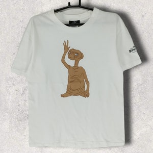 Puede incluir: Camiseta blanca con una ilustración de dibujos animados marrón de E.T. La figura extraterrestre levanta la mano en señal de saludo. La camiseta está colgada de una percha negra. La camiseta tiene un pequeño logotipo en la manga.