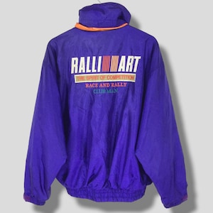 Puede incluir: Chaqueta Ralliart morada vintage con cuello alto y ribete naranja. La parte trasera de la chaqueta presenta el logotipo de Ralliart en blanco, rojo y naranja. También se puede ver el texto "The Spirit of Competition" y "Race and Rally Club Man".