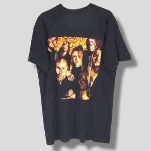 May include: Black t-shirt featuring a group portrait of musicians. The image on the shirt has a warm color palette with shades of yellow, orange, and brown. The t-shirt is a classic crew neck style.