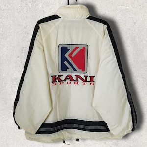 May include: Cream-colored Kani Sports jacket with black accents. The jacket features a large logo on the back with a red and blue K design and the word "KANI" in red. The jacket has a black stripe at the bottom and black trim along the sides.