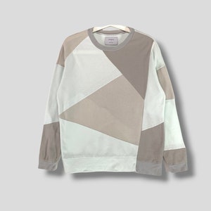 May include: A long-sleeved sweatshirt with a geometric patchwork design. The colour scheme includes light green, white, and various shades of brown. The sweatshirt is hanging on a white hanger.