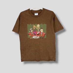Puede incluir: Camiseta marrón con una ilustración de dibujos animados de cuatro figuras en un sofá rojo, la palabra "NOBUST" en blanco. La camiseta tiene cuello redondo y mangas cortas. La camiseta está colgada de una percha blanca.