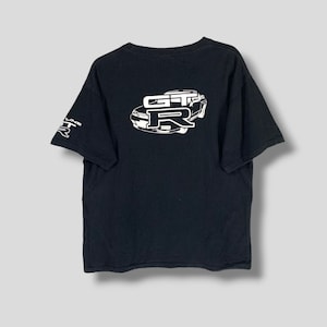 Puede incluir: Camiseta negra con un gráfico blanco de un coche con el texto "GT-R" en la espalda. La camiseta tiene un logotipo "GT-R" blanco en la manga izquierda.