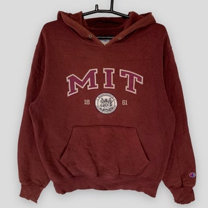 Puede incluir: Sudadera con capucha granate con las letras "MIT" en un diseño granate y blanco. La sudadera con capucha tiene un bolsillo delantero y una capucha. El logotipo de Champion está en la manga izquierda. La imagen también incluye los años 18 y 61, y un emblema circular.