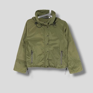 Puede incluir: Una chaqueta verde con capucha, cierre de cremallera y cordón de ajuste en la cintura. La chaqueta tiene dos bolsillos con cremallera en la parte delantera y un pequeño logotipo en el lado izquierdo del pecho.