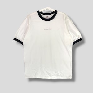 Puede incluir: Camiseta blanca con cuello redondo y ribetes negros en el cuello y las mangas. El texto "TOYOTA TEAM TUMI'S" está impreso en letras grises pequeñas. La camiseta está colgada en una percha blanca.