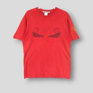 Puede incluir: Camiseta roja con un diseño gráfico en la parte delantera. El diseño presenta dos círculos y formas de alas. La camiseta tiene mangas cortas y cuello redondo. La camiseta está hecha de un material suave.