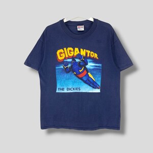 Puede incluir: Camiseta azul marino con un gráfico de Gigantor, un personaje robot, y el texto "GIGANTOR" en amarillo y rojo. El texto "THE DICKIES" está debajo. La camiseta está colgada en una percha blanca.