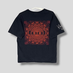 Puede incluir: Camiseta negra con un diseño geométrico rojo y naranja en la espalda, con la palabra "TOOL". La manga muestra texto blanco: "TOOL LOS ANGELES" y un símbolo de estrella.