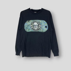 Puede incluir: Camiseta negra de manga larga con un gráfico de calavera y el texto "MEGADETH CRYPTIC WRITINGS" en forma de chapa de identificación. El gráfico es en tonos azules, verdes y blancos. La camiseta tiene cuello redondo y mangas largas.