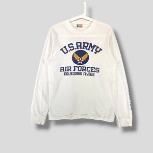 May include: White long-sleeve shirt with the text "U.S. ARMY AIR FORCES CALIFORNIA FLYERS" in navy blue. The shirt features a circular emblem with a star and wings. The shirt is hanging on a white hanger.