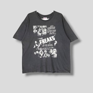 Puede incluir: Una camiseta negra con una impresión blanca de un cartel de película para la película de 1932 "The Freaks". El cartel presenta el texto "What Diabolical Vengeance Did These Half-Human Creatures Inflict Upon The Reality And The Strong Man?" y "The Most Amazing Cast Ever Assembled".