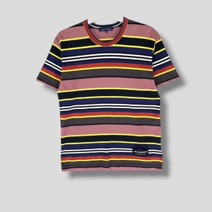 May include: A short-sleeved t-shirt with horizontal stripes in various colors, including pink, black, yellow, blue, red, and white. The shirt has a crew neck and a small label near the bottom. The shirt is hanging on a white hanger.
