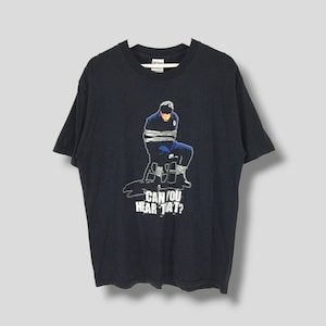 May include: Black t-shirt featuring a graphic of a person tied to a chair with the text "CAN YOU HEAR THAT?". The graphic is in shades of blue, white, and gray. The shirt is hanging on a white hanger.