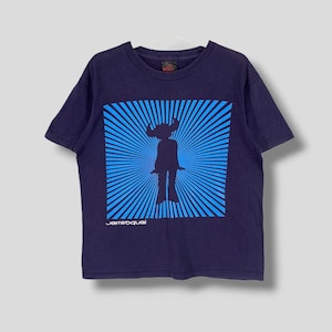 Puede incluir: Camiseta azul marino con un gráfico azul y celeste de una figura silueta con cuernos, sobre un diseño de rayos de sol. El texto "jamiroquai" está impreso debajo. La camiseta está colgada en una percha blanca.