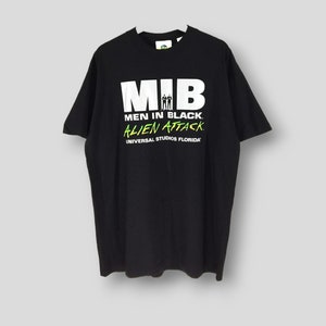 Puede incluir: Camiseta negra con el texto "MIB" en letras blancas grandes, y "MEN IN BLACK ALIEN ATTACK" debajo en blanco y verde. La camiseta también incluye "UNIVERSAL STUDIOS FLORIDA" en verde. Una camiseta clásica para los fans de la película.