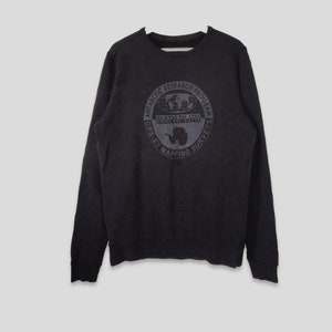 May include: Black crew neck sweatshirt with a circular graphic. The graphic includes the text "ANTARCTIC RESEARCH PROGRAM" and "NAPAPIJRI" with a map of the world and an outline of Antarctica. The sweatshirt is a solid black color.
