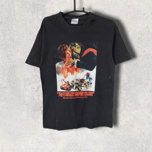 May include: Black t-shirt with a vintage movie poster design. The graphic shows a woman and a vampire, with the text "THE FEARLESS VAMPIRE KILLERS" and further text below. The shirt is suspended from a black hanger.