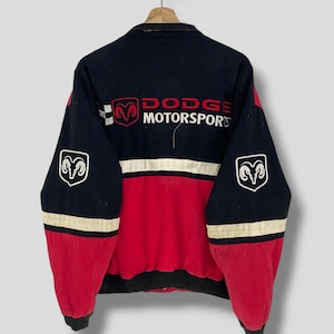 Op de afbeelding: Zwarte, rode en witte Dodge Motorsports jas. De jas heeft het Dodge-logo op de rug en mouwen, met de woorden "Dodge Motorsports" in het rood. De jas heeft een zwarte kraag en manchetten.