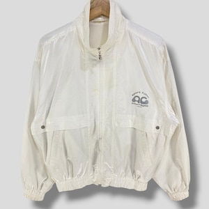 K&ouml;nnte beinhalten: Eine cremefarbene Vintage-Jacke mit hohem Kragen und durchgehendem Rei&szlig;verschluss. Die Jacke hat zwei Vordertaschen und ein Logo auf der rechten Brust mit der Aufschrift "sport futur" und "ac coutures". Die Jacke h&auml;ngt an einem Holzkleiderb&uuml;gel.