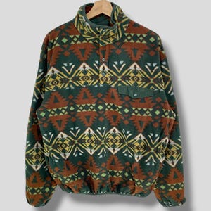 May include: A patterned fleece pullover in shades of green, brown, and yellow. The long-sleeved top features a high collar, a small chest pocket, and a button-up closure. The geometric design is repeated across the fabric.