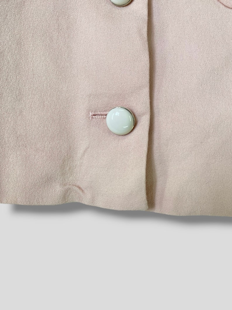 May include: A close-up of a light pink fabric with two white buttons. The buttons are sewn onto the fabric and have a silver rim.