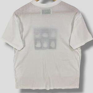 May include: White t-shirt with a crew neck and short sleeves. The back of the shirt features a rectangular graphic with a series of abstract shapes. The shirt is hanging on a wooden hanger.
