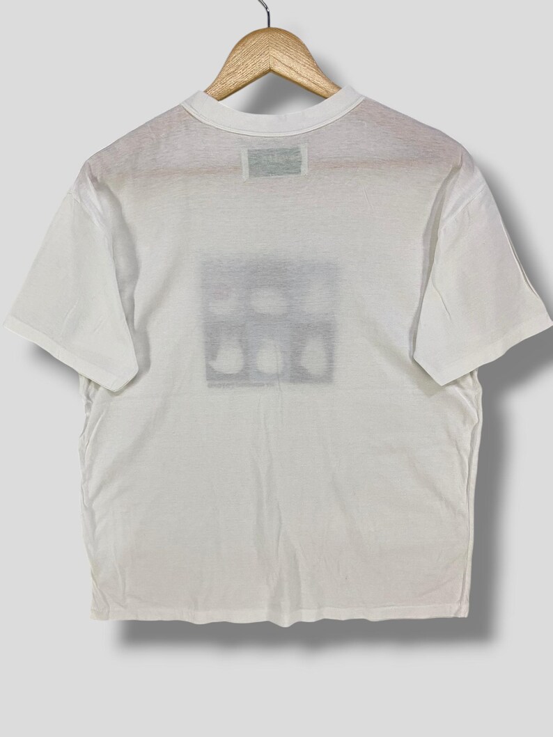 May include: White t-shirt with a crew neck and short sleeves. The back of the shirt features a rectangular graphic with a series of abstract shapes. The shirt is hanging on a wooden hanger.