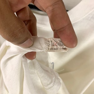 K&ouml;nnte beinhalten: Nahaufnahme eines wei&szlig;en T-Shirt-Kragens mit Etikett. Das Etikett enth&auml;lt Waschanweisungen und den Text "K-OS 5563 4". Das T-Shirt wird von einer Hand gehalten.