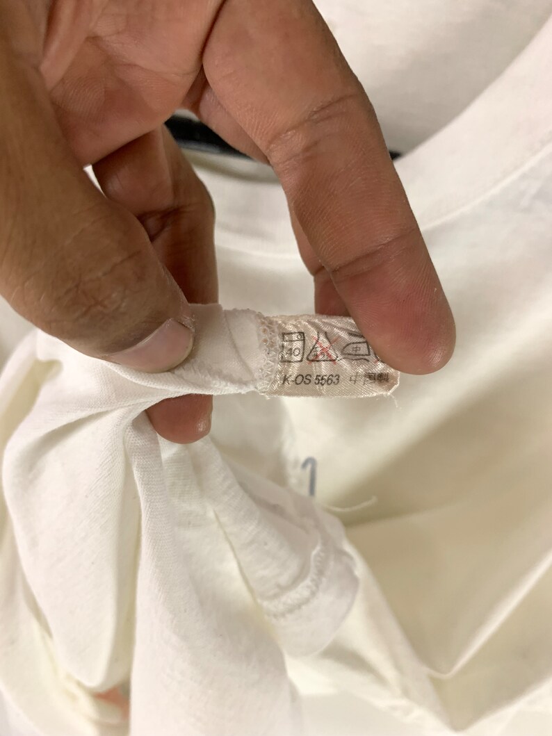 K&ouml;nnte beinhalten: Nahaufnahme eines wei&szlig;en T-Shirt-Kragens mit Etikett. Das Etikett enth&auml;lt Waschanweisungen und den Text "K-OS 5563 4". Das T-Shirt wird von einer Hand gehalten.