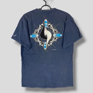 May include: Dark blue t-shirt with a graphic design on the back. The design features a stylized flame shape in white and black, set within a cross-like structure with blue accents. The shirt is hanging on a black hanger.