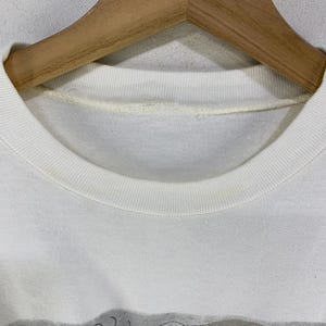 Puede incluir: Primer plano de una camiseta blanca con cuello redondo y un dise&ntilde;o gr&aacute;fico en la parte inferior. El gr&aacute;fico presenta formas abstractas en gris, morado y verde. La camiseta est&aacute; colgada en una percha de madera.