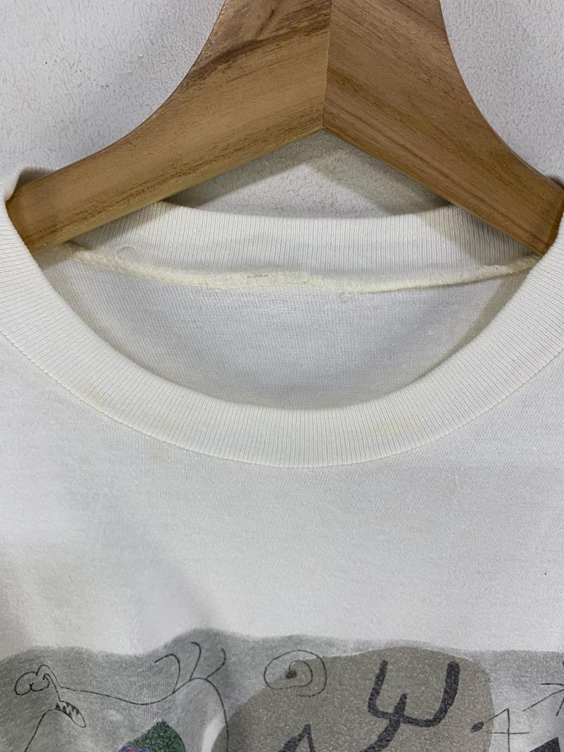 Puede incluir: Primer plano de una camiseta blanca con cuello redondo y un dise&ntilde;o gr&aacute;fico en la parte inferior. El gr&aacute;fico presenta formas abstractas en gris, morado y verde. La camiseta est&aacute; colgada en una percha de madera.